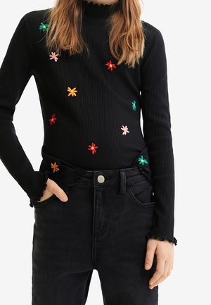 Haut noir côtelé à manches longues avec des fleurs brodées colorées porté par une personne ayant la main dans la poche avant d'un jean noir taille haute.