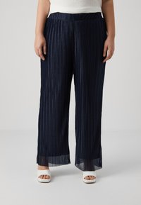 Navy pleated wide-leg pants with an elastic waistband, textured fabric, and sheer accents at the hem. Styled with white open-toe heels.