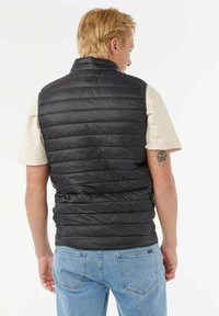 En blond mand vendt væk iført en sort quiltet vest over en beige kortærmet skjorte og lyseblå jeans, med en øjetatovering på sin arm.
