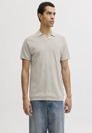 Άνδρας με κοντά σγουρά μαλλιά, ντυμένος με μπεζ πόλο με κοντά μανίκια και φαρδιά μπλε τζιν, στέκεται μπροστά σε απλό ανοιχτό φόντο.