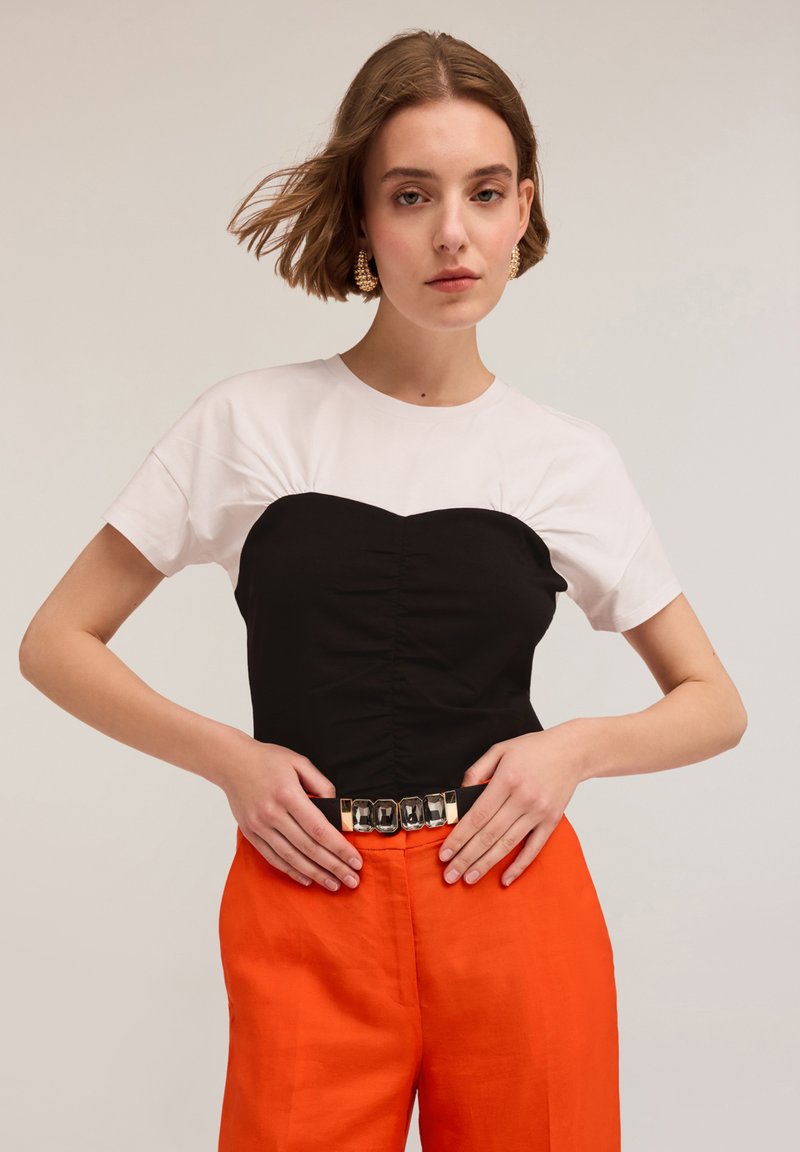 Top de manga corta en blanco y negro con un corsé negro ajustado, textura fruncida, combinado con pantalones naranjas brillantes y un cinturón con detalles en plata.