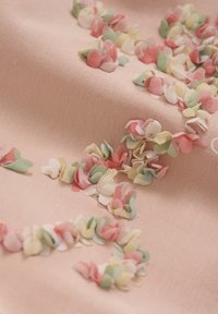 Textil med en mjuk rosa bakgrund, prydd med små tygblad i rosa, grönt och krämigt, med lager på lager av texturerade detaljer.