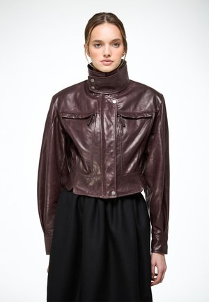 Jeune femme portant une veste en cuir marron foncé avec un col montant et des poches poitrine zippées, associée à une jupe noire, se tenant devant un fond blanc.