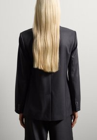 Personne avec de longs cheveux blonds raides portant une veste et un pantalon de costume noirs, se tenant contre un mur gris clair uni, dos à la caméra.