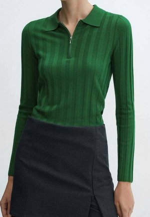 Kobieta ubrana w zielony prążkowany top z długim rękawem i półzamkiem przy kołnierzu, włożony do ciemnoszarej spódnicy z przednim rozcięciem.