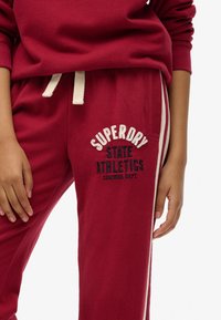 Røde bomulds sweatpants med cremefarvede side-striber. Har broderet branding "SUPERDRY STATE ATHLETICS" på låret. Elastisk talje med snoretræk.