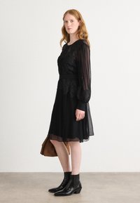 Robe noire avec des manches longues transparentes et des détails en dentelle florale. Jupe fluide, taille ceinturée, associée à des bottes noires et un sac marron.