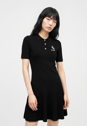 Femme aux longs cheveux noirs porte une robe polo noire à manches courtes avec des boutons blancs et un petit logo rose sur la poitrine.