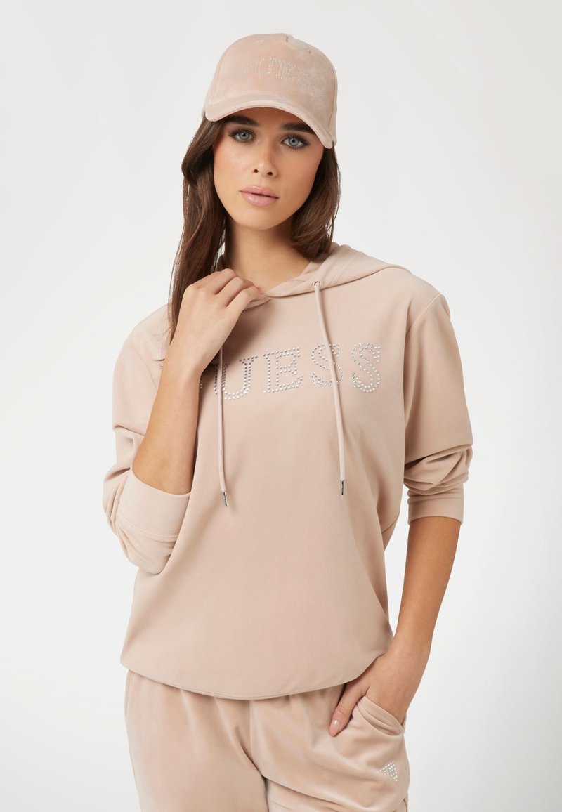 Felpa in velour rosa chiaro con lettering in strass "GUESS", cappuccio con cordino e vestibilità rilassata. Abbinata a pantaloni in velour coordinati e un berretto.