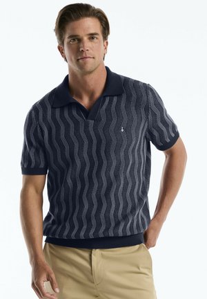 Mann trägt ein marineblau gemustertes kurzärmeliges Polohemd und beige Hose, steht mit einer Hand in der Tasche vor weißem Hintergrund.