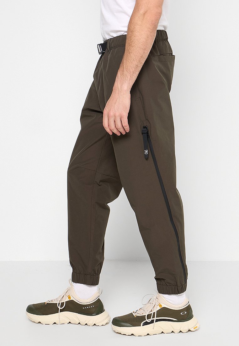 Pantaloni jogger verde oliva scuro con vita elasticizzata, polsini affusolati e tasca con zip. Tessuto liscio con una leggera lucentezza e vestibilità casual.