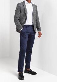 Blazer grigio a trama sopra una camicia bianca, abbinato a pantaloni blu scuro e mocassini neri. Il blazer presenta un singolo bottone e rever a punta.