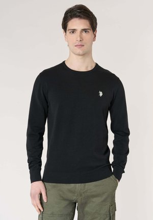 Giovane con capelli scuri che indossa una maglia nera a maniche lunghe con un piccolo emblema bianco e pantaloni cargo verde oliva, in piedi davanti a uno sfondo semplice.