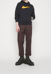 Svart hoodie med en mångfärgad Nike-logotyp, bruna byxor med uppvikta muddar och svarta stövlar med gul sömnad och snören.