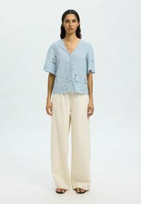Blusa azzurro chiaro con scollo a V e maniche corte a falbala. Presenta ricami ad occhiello e un orlo scallop. Abbinata a pantaloni larghi color crema.