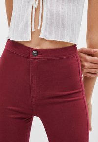 Parte media de una persona que lleva pantalones burdeos de cintura alta con cierre de botón y una blusa corta de punto blanca con lazos.