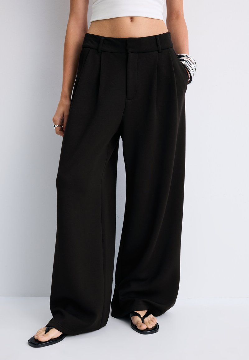 Person wearing loose black wide-leg pants, a white crop top, black flip-flops, and silver bangles on one wrist.