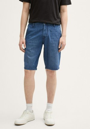 Jeansshorts in einem mittleren Blauton mit einer lockeren Passform, vorderen Taschen und einem umgeschlagenen Saum, kombiniert mit weißen Sportsneakern und Socken.