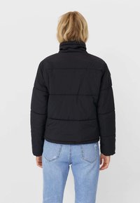 Zwarte pufferjack met een hoge kraag, horizontaal gestikt ontwerp en ribbelmanchetten, gecombineerd met lichtblauwe spijkerbroek.