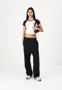 Kuttet hvit og svart T-skjorte med logo, kombinert med løse svarte bukser og lysfargede joggesko. Modellen holder en svart veske.