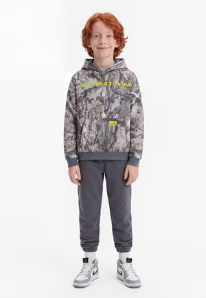 Enfant roux debout face à l’avant portant un sweat à capuche Jordan à motif camouflage, un pantalon de jogging gris, des chaussettes blanches et des baskets grises et blanches.