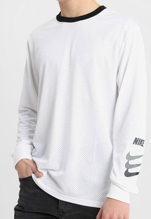 Osoba ubrana w białą, długorękawową koszulkę Nike z perforowaną tkaniną, czarnym kołnierzem oraz logo Nike na lewym rękawie.