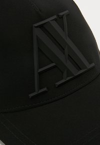 Casquette noire avec un logo "AX" en relief bien visible en noir mat, fabriquée en tissu avec une texture lisse et une visière courbée.
