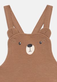 Salopette en coton brun avec un design de visage d'ours et des oreilles sur les bretelles, détail à boutons-pression et texture douce. Idéale pour les vêtements pour enfants.