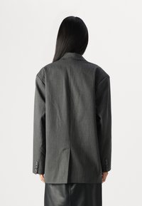 Grå pinstripet blazer med en løs passform, som har brede skuldre, lange ermer og knappe mansjetter, vist fra baksiden.
