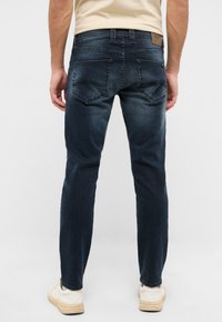 Vaqueros de mezclilla azul oscuro con un corte ajustado, que presentan dos bolsillos traseros con costuras decorativas y un parche de cuero en la cinturilla.