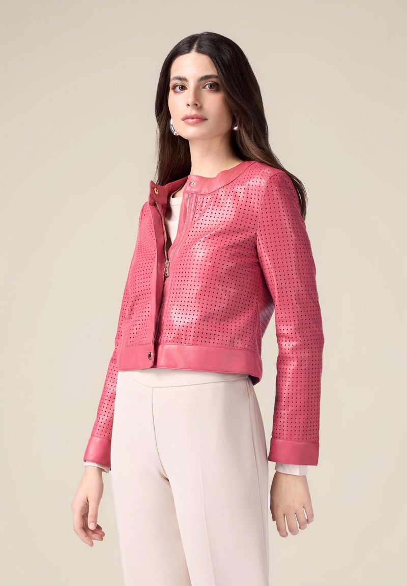 Giacca di pelle rosa tagliata con design perforato, chiusura con zip e polsini a contrasto. Abbinata a pantaloni beige chiaro.