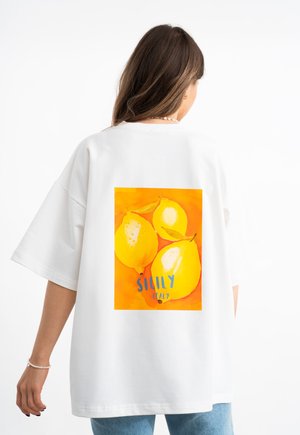 Witte oversized t-shirt met een rugafbeelding van drie gele citroenen op een oranje achtergrond, met het label "Sicilië Italië" in blauwe tekst.