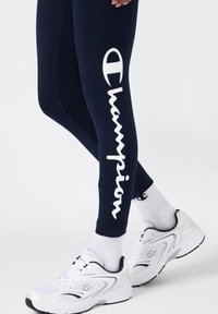 Granatowe legginsy z białym napisem i logo "Champion" na lewym udzie, zestawione z białymi butami sportowymi i szarymi skarpetami. Gładka tekstura materiału.