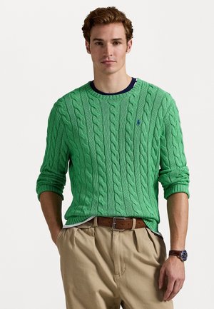 Suéter verde em malha trinada com punhos e gola canelados, combinado com calças bege e um cinto castanho. Relógio casual no pulso.