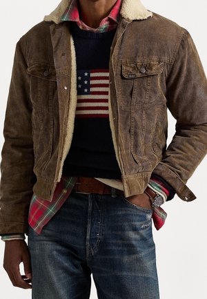 Mężczyzna ubrany w brązową kurtkę ze sztruksu z kołnierzem z owczej wełny, pod którą ma granatowy sweter z flagą amerykańską, czerwoną koszulę w kratę oraz niebieskie dżinsy.