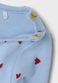Hellblaues Kleidungsstück mit roten Herzmustern, goldenen Druckknöpfen und glatter Baumwolltextur. Sichtbare Nähte an den Kanten.