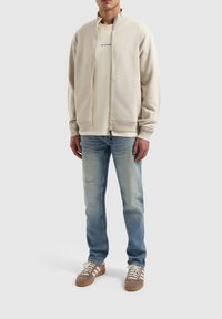 Beige zip-up sweatshirt over een licht shirt, gecombineerd met lichtblauwe jeans en bruine sneakers met witte strepen. Glad materiaal, casual pasvorm.