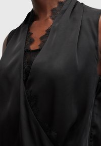 Gros plan sur un vêtement noir sans manches avec une bordure en dentelle le long du décolleté en V profond porté par une personne à la peau foncée.
