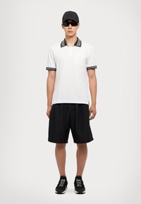 Biela polo košela s čiernym vzorovaným golierom, čierne šortky, čierne tenisky a slnečné okuliare, doplnené čiernou čiapkou s embosovaným textom.