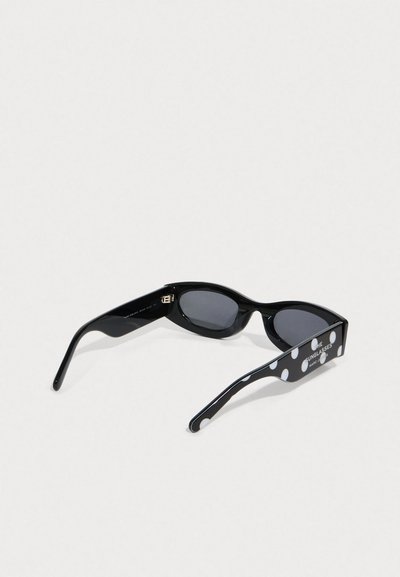 Gafas de sol negras con lentes oscuros en forma ovalada y varillas anchas, una de las varillas decorada con grandes lunares blancos y texto de marca.