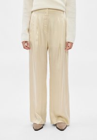 Άτομο που φοράει φαρδιά σατέν παντελόνια κρεμ χρώματος, μπεζ μυτερές μπότες και πλεκτό πουλόβερ κρεμ, στέκεται μπροστά σε απλό φόντο.