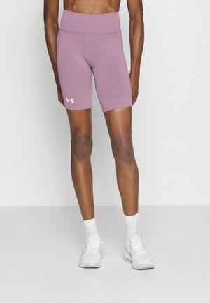 Personne portant un short de cyclisme mauve Under Armour et des chaussures de sport blanches avec des chaussettes, debout devant un fond clair uni.