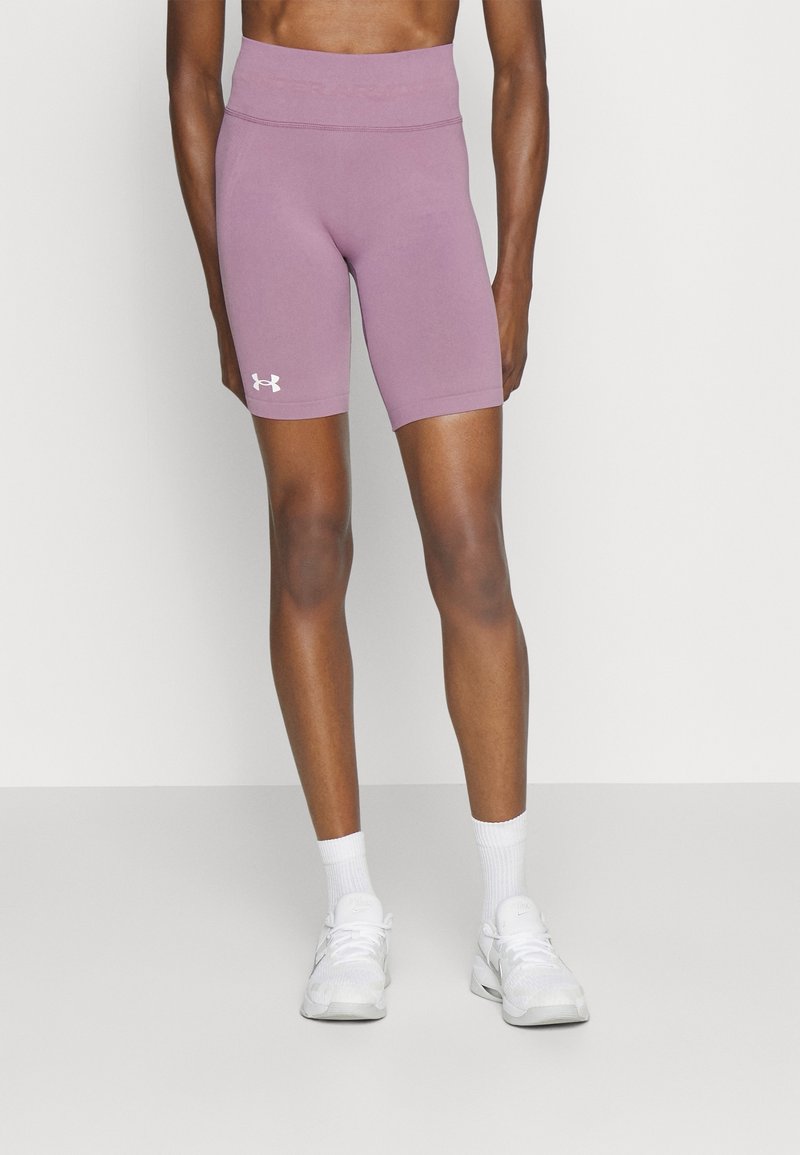 Personne portant un short de cyclisme mauve Under Armour et des chaussures de sport blanches avec des chaussettes, debout devant un fond clair uni.