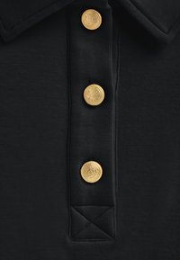 Polo nero con un tessuto testurizzato, tre bottoni rotondi dorati con design impressi, e un accento triangolare ricamato nella parte inferiore.