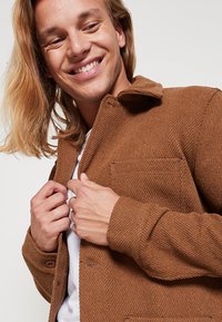 Brown textured jacket with a collar, button front, and chest pocket. Model wears a white shirt underneath, showcasing a casual style.