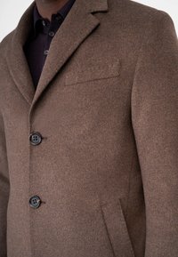 Brun ullkappa med notch-slagskrage, två framskjutna knappar och bröstficka. Har en mjuk textur och skräddarsydd passform, med sidofickor.