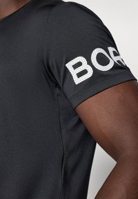 Schwarzes Sport-T-Shirt mit kurzen Ärmeln; zeigt auffälligen weißen "BORD"-Text auf dem Ärmel; glatte, enganliegende Stofftextur.