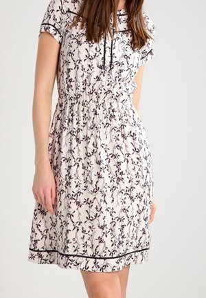 Woman wearing a white knee-length dress with black floral patterns, short sleeves, cinched waist, and black piping along the hem and neckline.