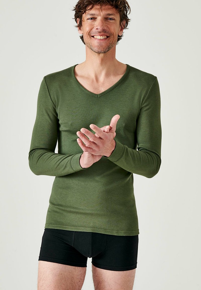 Langsleeve, V-hals thermoshirt in donkergroen, gemaakt van zachte stof, gedragen met zwarte boxers. Huid zichtbaar bij de hals en polsen.