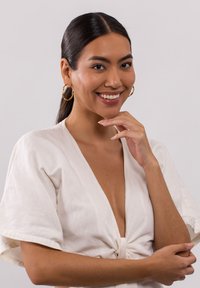 Weißes Leinentop mit kurzen Ärmeln und einem Knoten-Detail vorne. Das Model hat einen hellbraunen Hautton und trägt goldene Creolen.